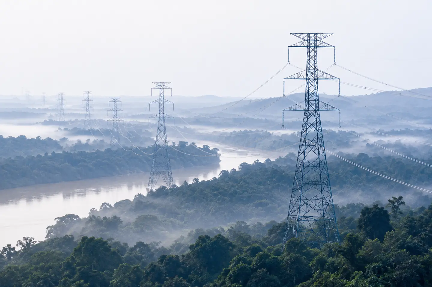 High-voltage transmission line crossing a forested river valley in morning mist